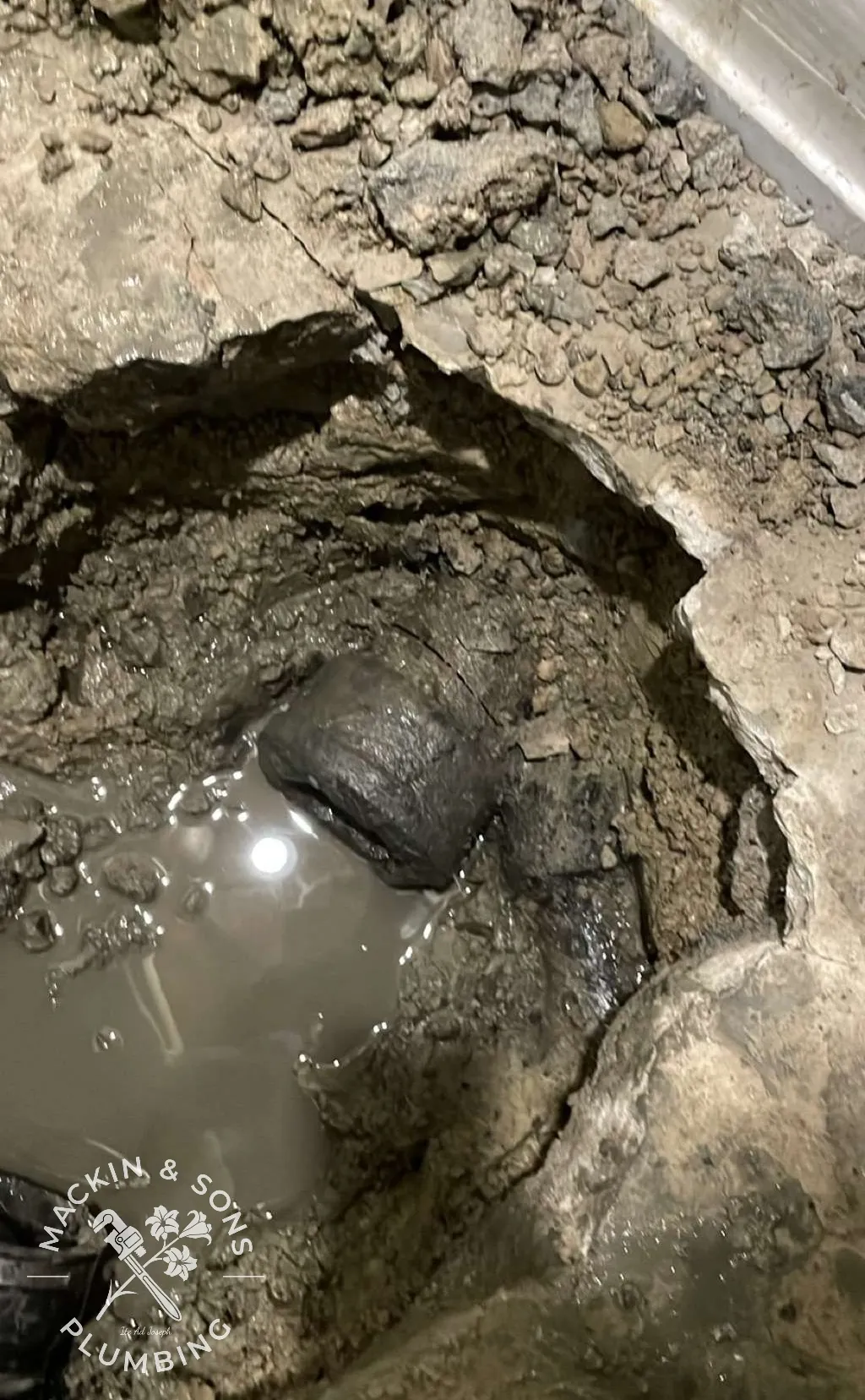Broken original cast iron drain pipe exposed in an excavated hole beneath a Medina basement floor, with muddy water pooling around it