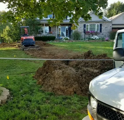 Mini excavator digging a sewer line trench across a front yard, with long piles of excavated soil and yellow utility marking flags along the lawn