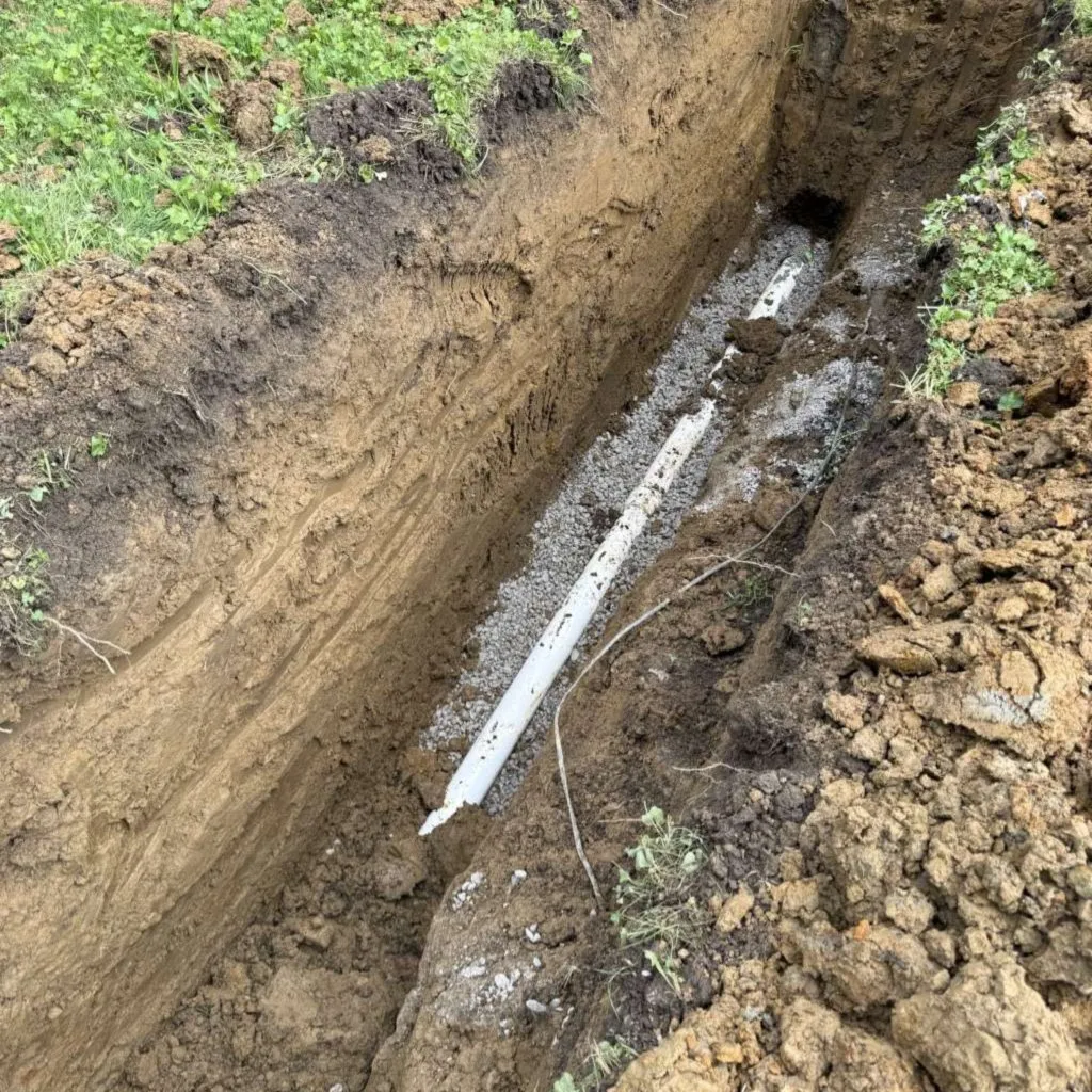 Open trench across a lawn with a new white PVC sewer pipe bedded in gravel at the bottom
