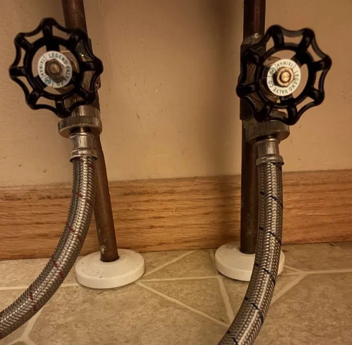 Two washing machine shut-off valves with braided stainless steel supply hoses at a laundry hookup